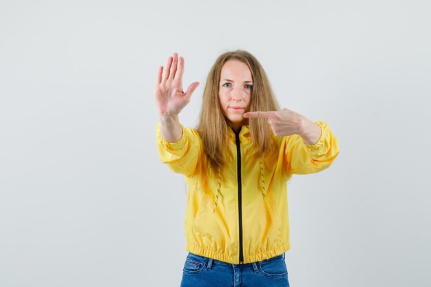 Young woman in yellow bomber jacket and blue jean showing stop gesture and pointing to it and looking serious , front view.