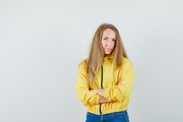 Young woman in yellow bomber jacket and blue jean standing with arms crossed and posing at camera and looking charming , front view.