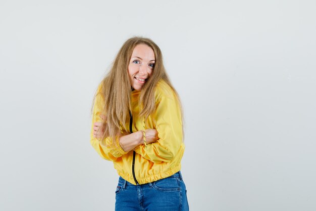 Young woman in yellow bomber jacket and blue jean standing with arms crossed and posing at camera and looking optimistic , front view.