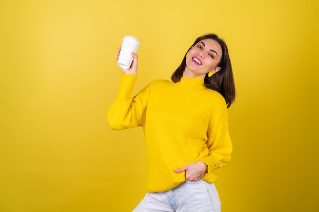 Giovane donna in un maglione giallo accogliente con lucidalabbra rosa brillante con una tazza di carta di caffè aromatico caldo, aspetto sognante, sorriso carino, calore radiante