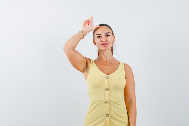 Young woman in yellow dress showing loser sign over head and looking pensive , front view.