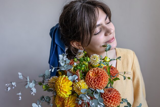 彼女の髪に青いリボン、黄色とオレンジ色の菊の花束、灰色の背景を持つ黄色のドレスを着た若い女性。