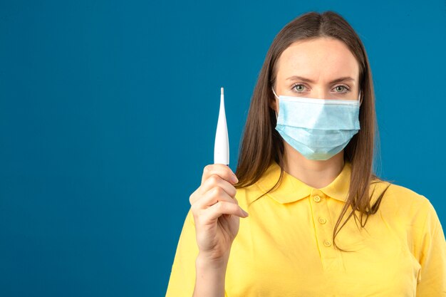 Young woman in yellow polo shirt and medical protective mask holding thermometer in hand looking at camera with serious face on isolated blue background
