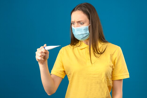 Young woman in yellow polo shirt and medical protective mask looking at thermometer in panic on isolated blue background
