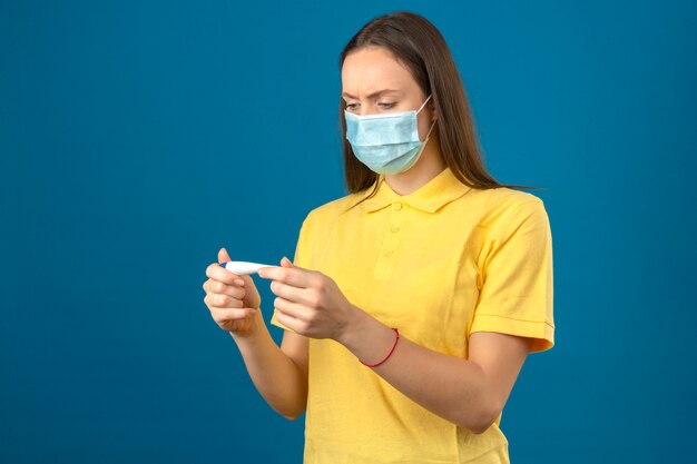 Young woman in yellow polo shirt and medical protective mask looking at thermometer with serious face on isolated blue background