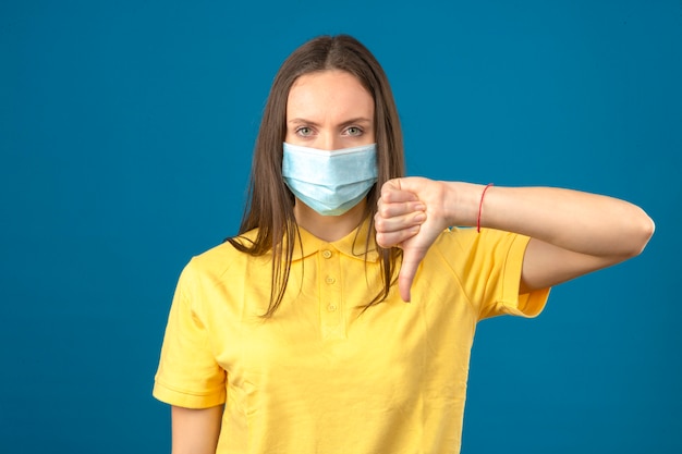 Молодая женщина в желтой рубашке поло и медицинской защитной маске, делая пальцы вниз знак серьезно глядя на камеру на изолированных синем фоне