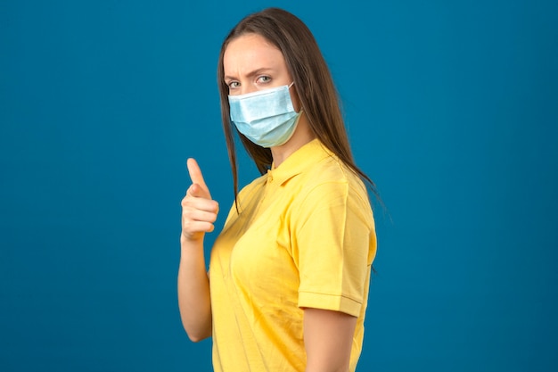 노란색 폴로 셔츠와 의료 보호 마스크 파란색 격리 된 배경에 서 심각한 얼굴로 카메라에 손가락을 가리키는 젊은 여자