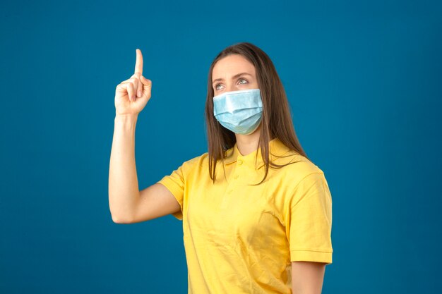 노란색 폴로 셔츠와 의료 보호 마스크 격리 된 파란색 배경에 손가락을 가리키는 젊은 여자