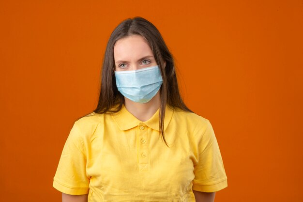 Young woman in yellow polo shirt and medical protective mask standing on orange background