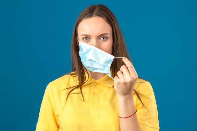 Молодая женщина в желтой рубашке поло, снимая медицинскую защитную маску, глядя на камеру с серьезным лицом на изолированных синем фоне