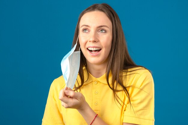 Young woman in yellow polo shirt taking off medical protective mask smiling with happy face isolated on blue background