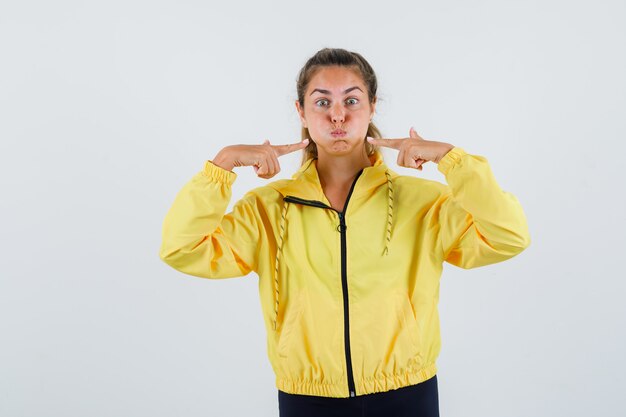 Young woman in yellow raincoat pointing at her puffed cheeks