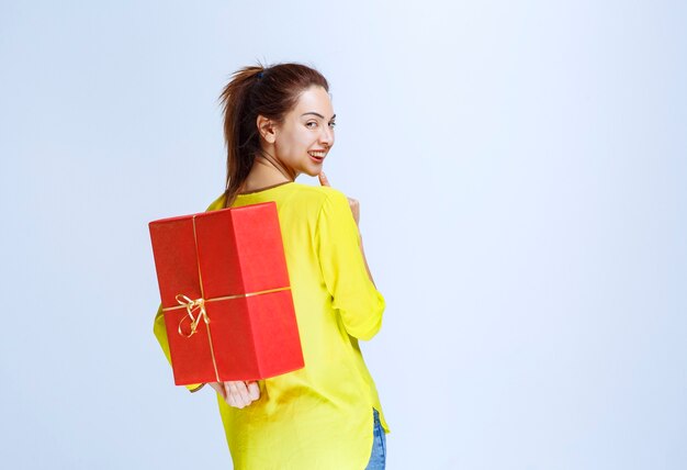 Young woman in yellow shirt hiding a red gift box behind herself