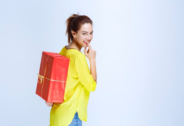 Молодая женщина в желтой рубашке прячет за собой красную подарочную коробку