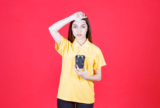 Young woman in yellow shirt holding a black disposable coffee cup and looks tired and sleepy