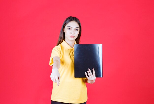 Young woman in yellow shirt holding a black folder, pointing and calling her colleague