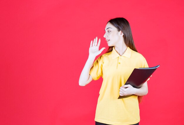 Молодая женщина в желтой рубашке держит черную папку, указывая и зовет своего коллегу