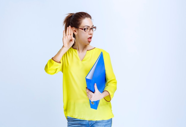 Young woman in yellow shirt holding a blue folder and looks confused and surprized