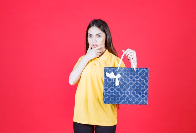 Young woman in yellow shirt holding a blue shopping bag and looks confused and thoughtful