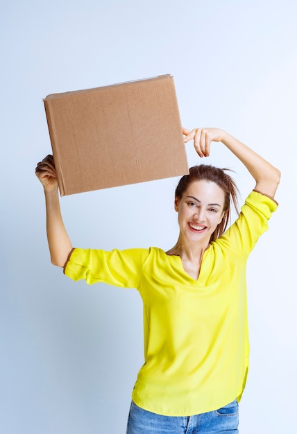 Молодая женщина в желтой рубашке держит картонную грузовую коробку и чувствует себя счастливой