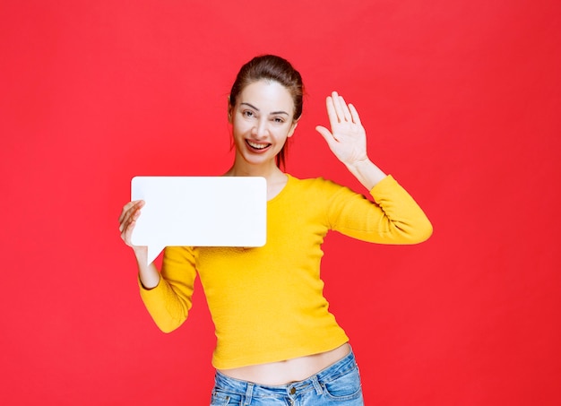 Young woman in yellow shirt holding a rectangle info board and greeting someone