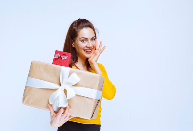 Young woman in yellow shirt holding a red and a cardboard gift boxes and getting surprised and thoughtful