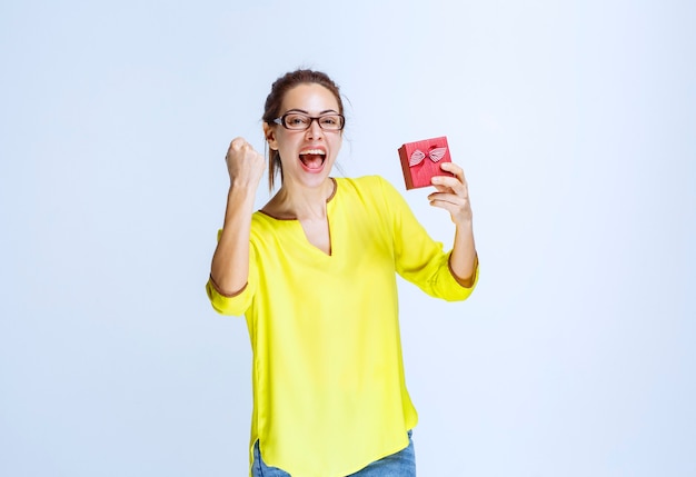 Young woman in yellow shirt holding a red gift box and showing enjoyment hand sign
