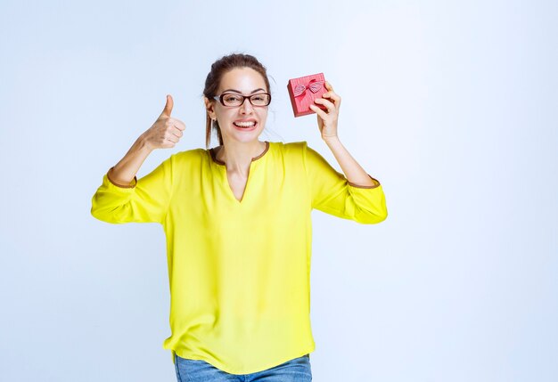 Young woman in yellow shirt holding a red gift box and showing enjoyment hand sign