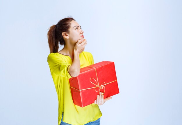 Young woman in yellow shirt holding a red gift box and thinking or hesitating