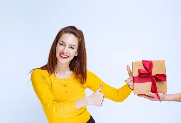 Молодой женщине в желтой рубашке предлагают картонную подарочную коробку с красной лентой и положительным знаком рукой