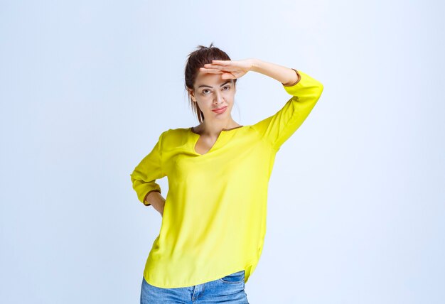 Молодая женщина в желтой рубашке, прикладывая руку ко лбу и наблюдая впереди