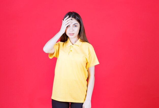 빨간 벽에 서서 피곤하고 졸려 보이는 노란 셔츠를 입은 젊은 여성