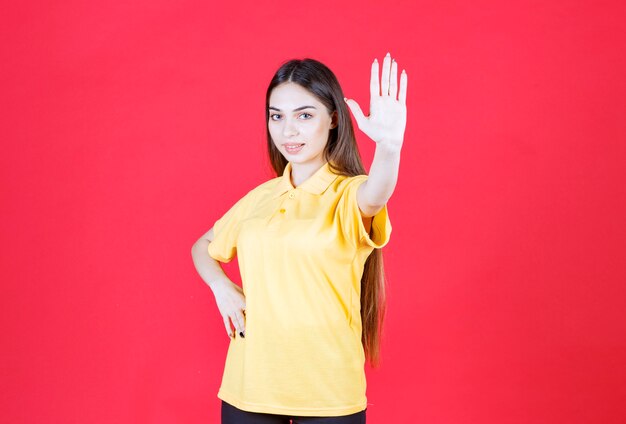 Молодая женщина в желтой рубашке стоит на красной стене и что-то останавливает