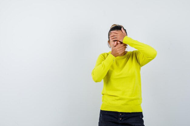 Young woman in yellow sweater and black pants covering face with hands and looking shocked 