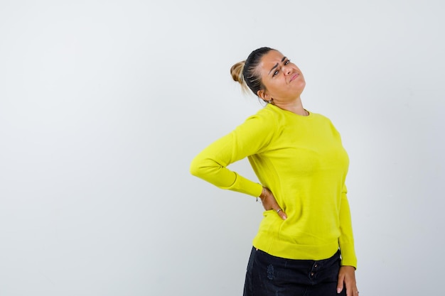 Foto gratuita giovane donna in maglione giallo e pantaloni neri che tiene la mano dietro la vita e sembra tormentata