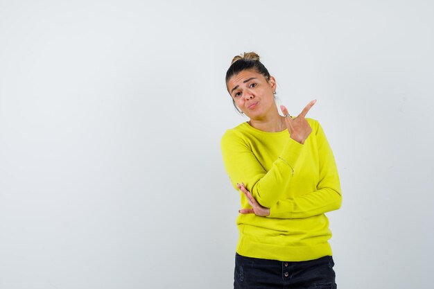 Young woman in yellow sweater and black pants pointing right while holding hand on elbow and looking pensive