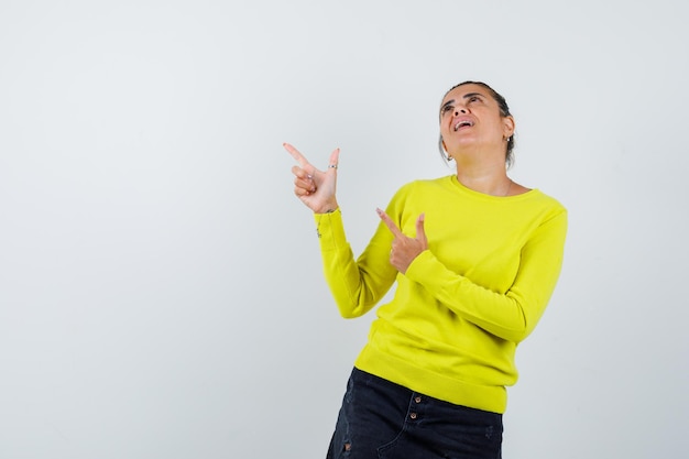 Young woman in yellow sweater and black pants pointing up, looking above and looking happy 