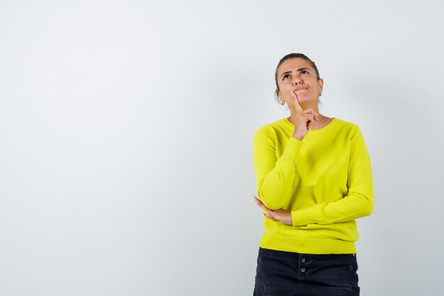 Young woman in yellow sweater and black pants putting index finger near mouth, thinking about something and looking pensive 