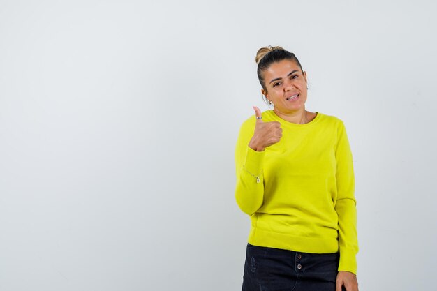 Young woman in yellow sweater and black pants showing thumb up and looking happy