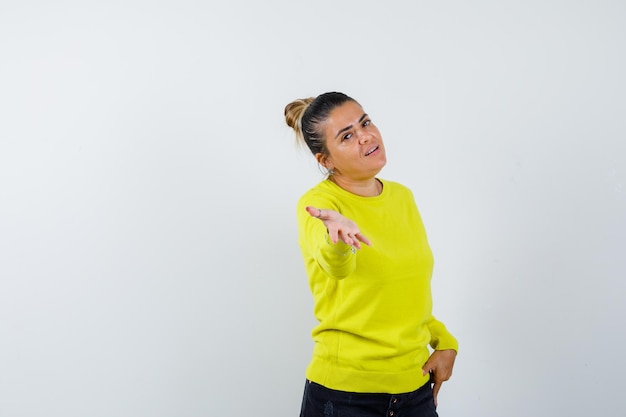 Young woman in yellow sweater and black pants stretching hand toward camera and looking happy 