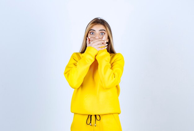 Young woman in yellow sweatsuit covering her mouth over white wall