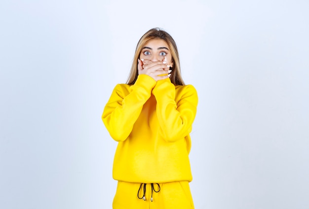 Young woman in yellow sweatsuit covering her mouth over white wall