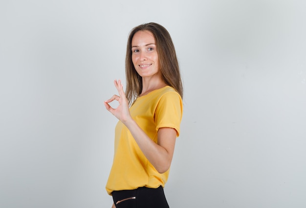 Young woman in yellow t-shirt, black pants showing ok gesture and looking pleased