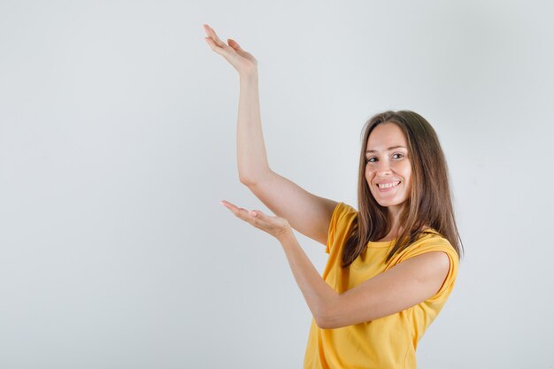 Молодая женщина в желтой футболке держит руки, будто поднимает что-то, и выглядит веселой