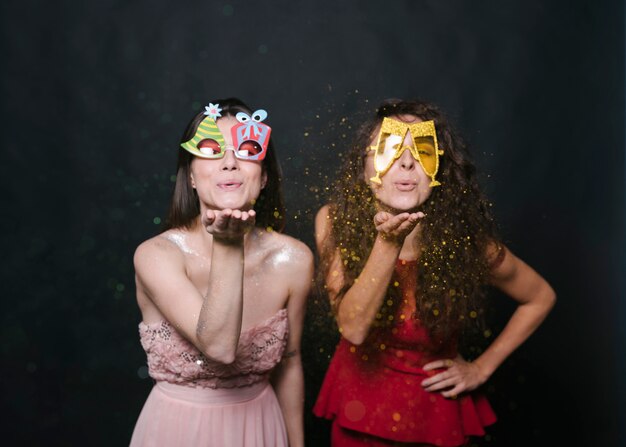 Young women in funny eyeglasses blowing confetti from hands