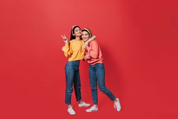 Young women in great mood show peace sign and pose on red wall