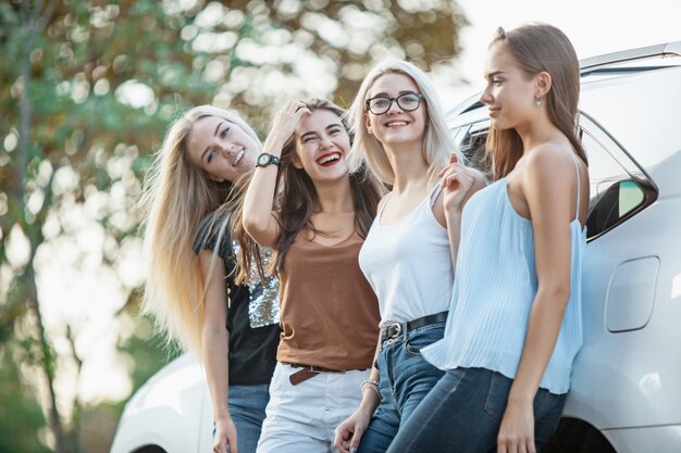 The young women standing and smiling near the car outdoor. The lifestyle, travel, adventure and female friendship concept