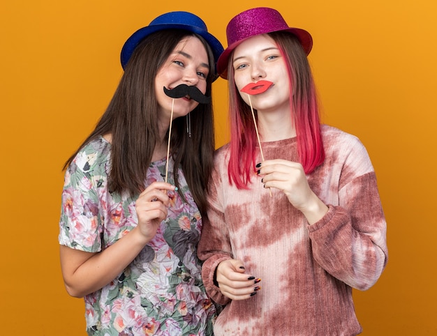 Giovani donne che indossano cappello da festa che tengono baffi finti su un bastone isolato su una parete arancione