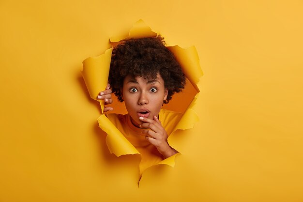 Young wondered young woman with Afro hairstyle, opens mouth, stands in ripped paper hole background