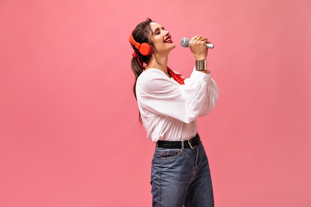 Молодая замечательная женщина в хорошем настроении поет в микрофон и слушает музыку в наушниках на розовом изолированном фоне.